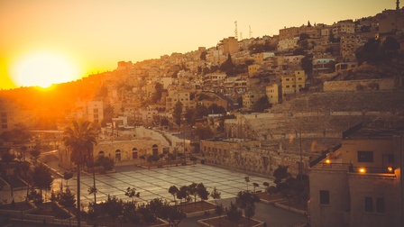 Panoramaaufnahme von Amman mit Wohnhäusern, einem Platz und Palmen. Am Horizont geht die Sonne auf