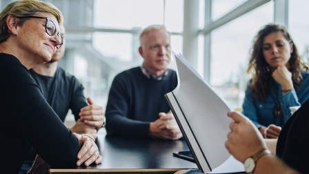 Mehrere Personen sitzen an Konferenztisch, Ausschnitt einer Person, die in Dokumentenmappe blättert 