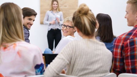 Rückenansicht, sechs neben- und hintereinander sitzender Personen, die teilweise nach vorne blicken und sich gegenseitig anblicken. Ihnen gegenüber steht eine weitere Person vor einer Wand, auf der ein großes, braunes Stück Papier hängt