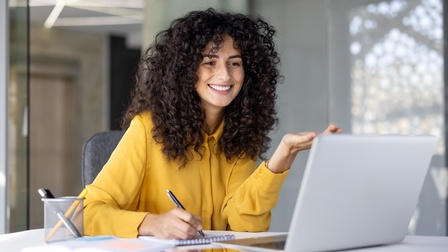 Eine Frau sitzt in einem Büro vor einem Laptop an einem Schreibtisch. Die Person notiert etwas mit einem Stift auf einem Notizblock und blickt auf den Bildschirm. Neben dem Laptop sind ein aufgeschlagenes Buch und ein Tablet