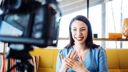 Lächelnde Person mit dunklen Haaren und roten Lippen strahlt in die Kamera, die im Vordergrund unscharf in Bild kragt