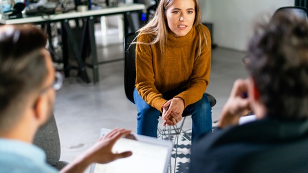 Person mit braunem Pullover und langen blonden Haaren sitzt auf einem Sessel und spricht zu anderen Personen, die ihr in der Unschärfe gegenübersitzen