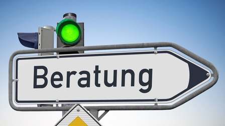 Auf einem Verkehrsschild, das nach rechts deutet, steht Beratung. Dahinter ist eine Ampel mit einem grünen Licht. Hinter den Verkehrszeichen ist der blaue Himmel
