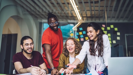 Personen arbeiten gemeinsam an einem Schreibtisch und blicken dabei auf ein Flipchart in einem modernen Arbeitsraum mit Glastrennwänden und Post-its während im Hintergrund ein Flur ersichtlich ist