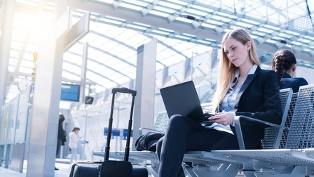 Person sitzt auf Sitzgelegenheiten in Bahnterminal und hat aufgeklapptes Notebook am Schoß, nebenstehend Rollkoffer
