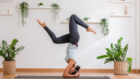 Seitliche Aufnahme einer Person in Sportkleidung auf einer Sportmatte in einem hellen Raum mit Topfpflanzen. Die Person macht einen Kopfstand. Sie hat die Hände auf dem Hinterkopf und die Beine angewinkelt