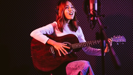 Eine Person sitzt vor einer dunklen Wand auf einem Hocker. Sie hält eine Gitarre in den Händen und spielt darauf. Vor ihr steht ein Mikrofon, in das die Person singt. Auf dem Kopf trägt sie Kopfhörer