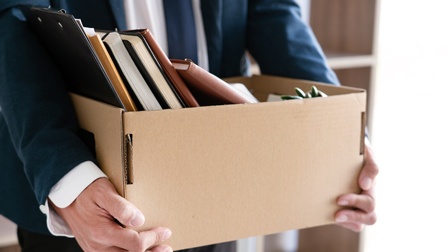 Nahaufnahme eines kleinen, rechteckigen Kartons, der von den Händen einer Person in einem Anzug gehalten wird. In dem Karton sind Bücher, Notizhefte und ein Klemmbrett vertikal nebeneinander angeordnet.