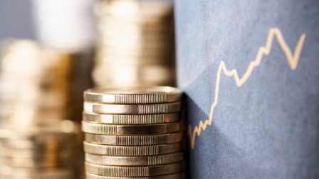 Vier Stapel Münzen sind neben einem grauen Display, auf dem ein Aktienkurs abgebildet ist