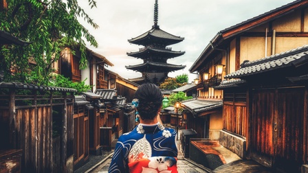 Rückenansicht einer Person in einem blau-weiß-rotem Oberteil mit großen Blumen sowie einer hochgesteckten Frisur, die auf einer Straße durch einen Ort geht. Links und rechts sind einstöckige Gebäude