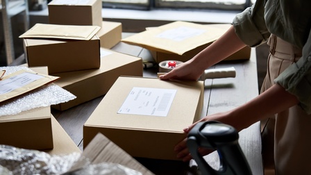 Auf einem Tisch liegen mehrere Pakete, Kuverts sowie durchsichtige Folie. Eine Person steht seitlich vor dem Tisch und hat ihre beiden Händen auf einem Paket