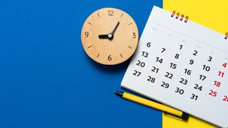 Auf blau-gelbem Hintergrund liegt eine Uhr aus Holz. Neben der Uhr liegt ein Kalenderblatt eines Ringbuchkalenders, unter dem ein Kugelschreiber liegt