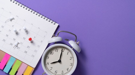 Ein Wecker liegt mit einem Wochenkalender, Pinnnadeln und Pos-its auf einem violetten Untergrund