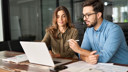 Nahaufnahme von Personen in einem Büro, hinter ihnen ist eine Glaswand, sie betrachten einen Laptop, eine Person hält einen Stift. Auf einem braunen Holztisch mit Maserung liegen Unterlagen und Notizbücher.