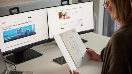 Person mit Brille sitzt an einem Schreibtisch und blickt auf eine Mappe mit Prüfungszeugnis Meisterprüfung, die sie in Händen hält, vor ihr stehen zwei Monitore, ein Laptop sowie eine Tastatur am Tisch