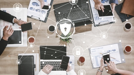 Top-Shot einer hölzernen Oberfläche, auf der aufgeklappte Laptops, Klemmbretter mit Papieren mit Statistiken, Tassen mit Flüssigkeiten, die Hände von Personen mit Smartphones sind. Darüber sind kleine Schlösser umgeben von Kreisen