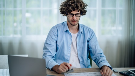 Person mit Brillen in blauem Hemd sitzt an Schreibtisch vor aufgeklapptem Laptop und stempelt vor sich liegenden Plan
