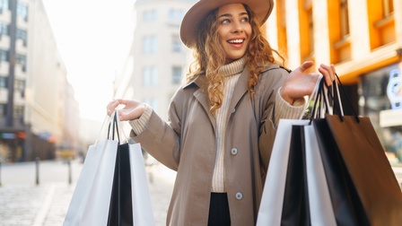 Eine Person mit Mantel und Hut steht in einer Fußgängerzone und hält mit beiden Händen mehrere Einkaufstüten. Im Hintergrund sind verschiedene Gebäude