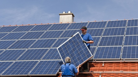 Zwei Personen hieven ein Solarpanel auf Hausdach, auf dem bereits andere Panele montiert sind, unter blauem Himmel