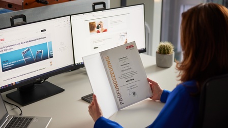 Person mit langen braunen Haaren und blauer Bluse sitzt an einem Schreibtisch mit Laptop und zwei Monitoren und hält ein Dokument mit Aufschrift Prüfungszeugnis Meisterin in Händen