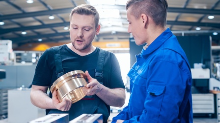 Zwei Personen in Overalls im Werkshalle stehend im Gespräch miteinander: Eine Person hält breites metallenes Verbundstück in Händen und blickt darauf sprechend