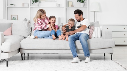 Zwei erwachsene Personen und zwei Kinder sitzen auf einer grauen Couch und spielen, im Hintergrund zeigt sich eine Wohnzimmer-Situation