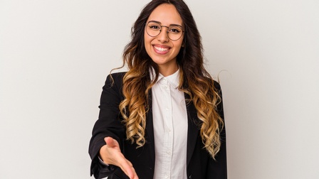 Porträt einer lächelnden Person mit offenen, lockigen Haaren, Brille, weißer Bluse und dunklem Sakko, die ihre rechte Hand in Richtung Kamera streckt