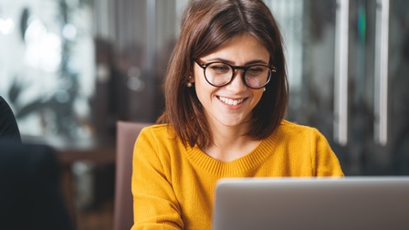 Portrait von einer lächelnden Person mit dunklen schulterlangen Haaren und Brille sowie gelbem Pullover, die vor einem Laptop sitzt