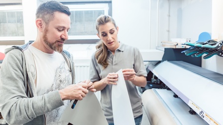 Zwei Personen lösen Klebefolie von Papierstreifen vor Beschriftungsmaschine stehend