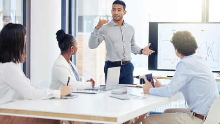 An einem Tisch sitzen drei Personen in Bürokleidung. Neben dem Tisch steht eine weitere Person, die zu den anderen spricht. Neben ihr ist ein großer Bildschirm, auf dem Statistikkurven sind
