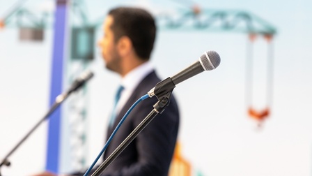 Seitliche Aufnahme eines Mikrofons, das an einem Mikrofonständer fixiert ist, im Vordergrund. Daneben steht eine Person  in einem Sakko mit kurzen Haaren, die in ein Mikrofon spricht, das vor ihr steht