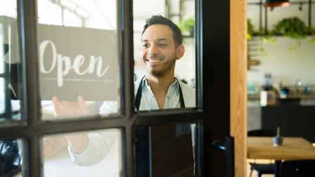 Lächelnde Person mit Schürze lugt durch Lokaltür und greift nach Schild auf dem Open zu lesen ist