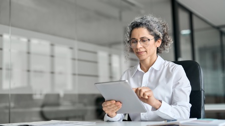 Eine Person mit Brille und weißem Hemd sitzt an einem Tisch. In den Händen hält sie ein Tablet, auf das sie blickt. Auf dem Tisch sind verschiedene Unterlagen und ein aufgeklapptes Buch. Links neben der Person ist eine Glaswand