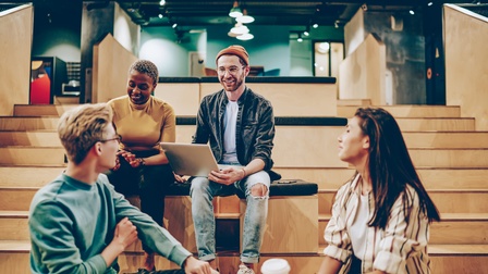 Vier Personen sitzen in zwei Reihen auf Holztreppen in einem Raum. Die Personen, die auf den unteren Treppen sitzen, haben sich zu den oberen zugewandt. Eine Person hat einen aufgeklappten Laptop auf dem Schoß