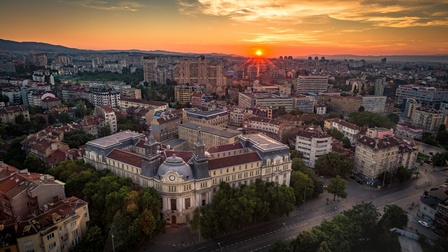 Panoramaaufnahme eines Stadtteils von Sofia mit verschiedenen Gebäuden aus unterschiedlichen Epochen, Straßen und Bäumen. Am Horizont steht die Sonne tief
