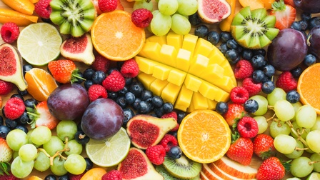 Topshot verschiedener Lebensmittel wie Erdbeeren, Himbeeren, Weintrauben, einer Mango, Feigen, Limetten, Pflaumen, Heidelbeere, Äpfel, Kiwis, Orangen.