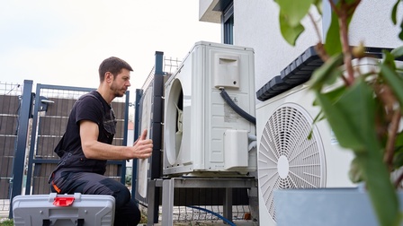 Eine Person installiert kniend eine Wärmepumpe an der Außenwand eines Hauses.