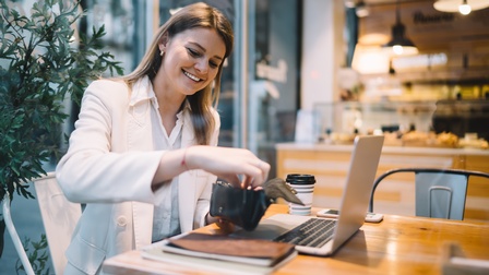Seitliche Aufnahme einer Person, die an einem Tisch in einem Lokal sitzt. Vor ihr sind ein aufgeklappter Laptop, ein Notizbuch, ein Kaffeebecher und ein Smartphone. Die Person holt aus ihrer Geldtasche Geld. 