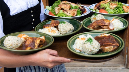 Nahaufnahme eines Tabletts, das von einer Person getragen wird. Auf dem Tablett sind sechs Teller, auf denen Speise liegen, zum Beispiel mit Knödeln und Fleisch oder mit grünem Salat und Fleisch.
