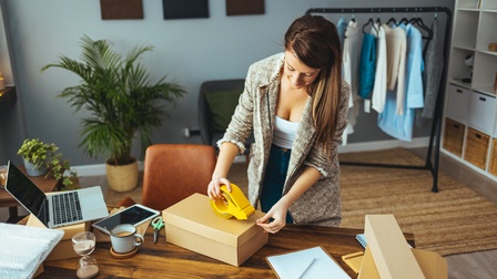 Person mit langen braunen Haaren verpackt eine Kartonage mit Klebeband bei einem Schreibtisch, im Hintergrund steht eine Kleiderstange mit Kleidung in einem Büroraum