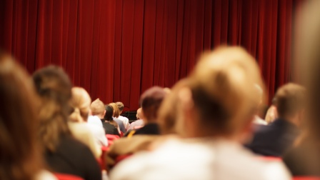 Das Publikum im Theater blickt auf den geschlossenen, roten Vorhang auf der Bühne.