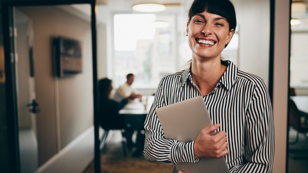 Porträt einer lächelnden Person mit dunklen Stirnfransen und gestreifter Bluse, die in einem Flur steht und mit beiden Händen einen Laptop beim Körper hält, im Hintergrund zeigen sich helle Meetingräume