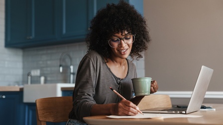 Eine Person mit Brille und dunklen lockigen Haaren sitzt an einem Holztisch in einer Küche. In der linken Hand hält sie einen Bleistift, mit dem sie etwas auf einem Blatt Papier notiert. Vor ihr steht ein aufgeklappter Laptop