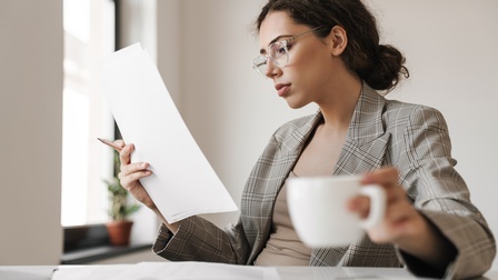 Person mit Brillen an Tisch sitzend hält Tasse in Hand und Dokument, auf das sie blickt