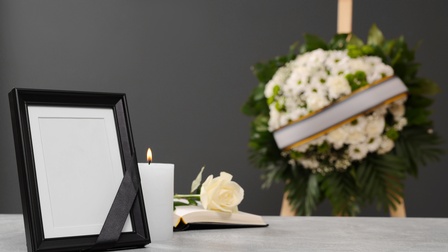 Schwarzer Stahbilderrahmen mit schwarzem Band auf Tisch stehend, daneben brennende weiße Kerze und aufgeschlagenes Buch mit weißer Rose, im Hintergrund verschwommen weißgrünes Blumenbouquet mit Scherpe