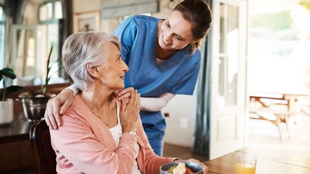 Person mittleren Alters mit kurzen grauen Haaren sitzt bei einem Tisch, vor ihr steht eine Schüssel sowie ein Glas, dahinter steht eine Person in blauer Arbeitskleidung, die wohlwollend die Hände auf ihren Schultern platziert