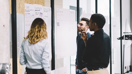 An eine Glaswand sind zwei große Zettel geklebt, auf denen Wörter wie Übersicht oder Email stehen. Vor den Zetteln stehen drei Personen, die darauf blicken