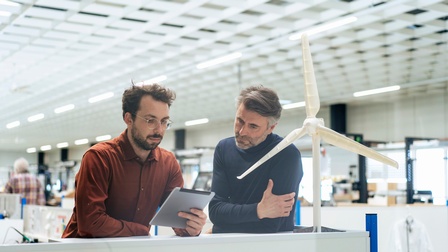 Personen blicken auf ein Tablet und lehnen sich an eine Front auf dem ein Modell einer Windturbine in einem Werksraum steht