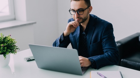 Person mit Brillen blickt auf aufgeklappten Laptop, eine Hand ans Kinn gestützt