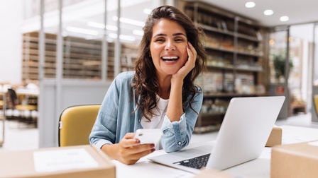 Lachende Person vor Laptop in Großraumbüro sitzend hält in einer Hand ein Smartphone, die andere Hand den Kopf stützend, ringsum Kartonagen